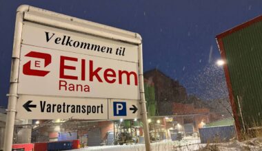 Skiltet viser "Velkommen til Elkem Rana" med retninger til Varetransport og parkering. Omgivelsene er snødekte med bygninger i bakgrunnen. Det er kveldstid, og snø faller mens lysene i anlegget lyser opp. Bildet gir inntrykk av industriområde i vintervær.