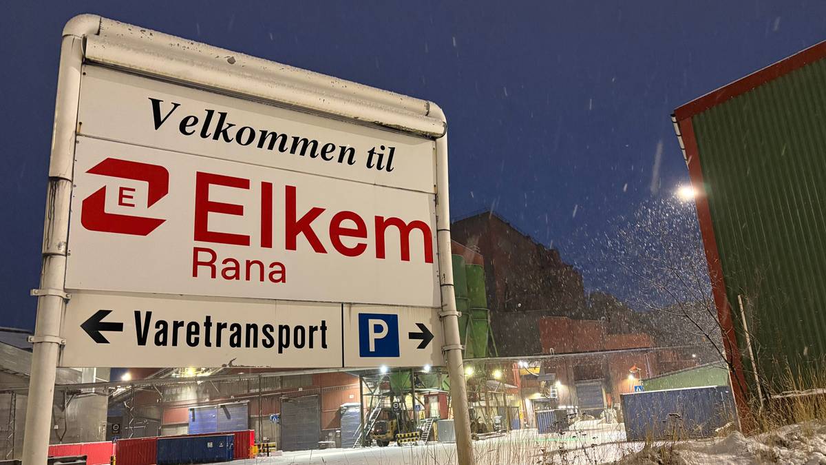 Skiltet viser "Velkommen til Elkem Rana" med retninger til Varetransport og parkering. Omgivelsene er snødekte med bygninger i bakgrunnen. Det er kveldstid, og snø faller mens lysene i anlegget lyser opp. Bildet gir inntrykk av industriområde i vintervær.