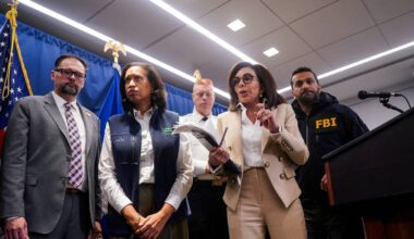 Flere personer står samlet foran en blå bakgrunn med amerikanske flagg. En kvinne i beige dress holder en gjenstand mens hun snakker. Andre personer er til stede, iført formelle klær og FBI-uniform. Det er en mikrofonscene i forgrunnen, trolig fra en pressekonferanse.