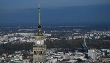 Utsikt over Warszawas skyline med Kultur- og vitenskapspaladset i fokus. Klokken på bygningen viser omtrent 11:00. I bakgrunnen er det flere moderne bygninger og elven Wisła. Bilde tatt på en klar dag med minimal skydekke.