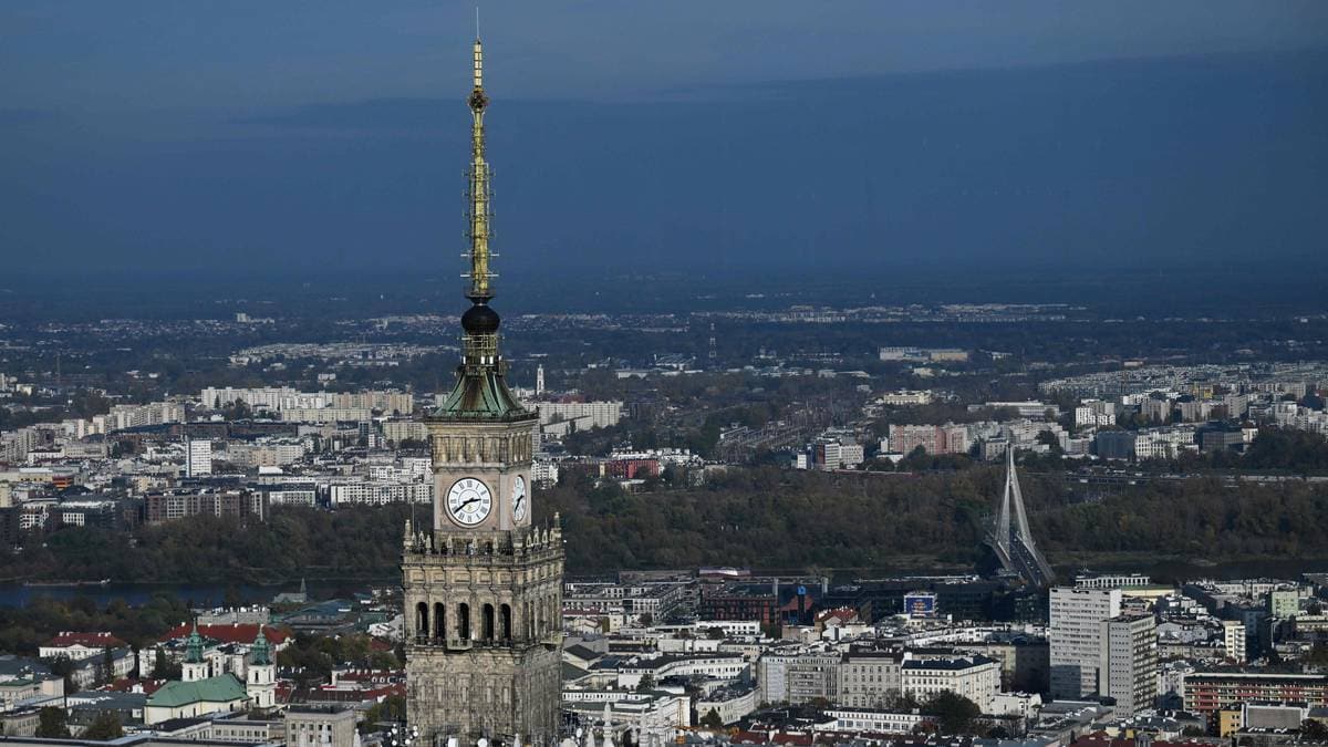 Utsikt over Warszawas skyline med Kultur- og vitenskapspaladset i fokus. Klokken på bygningen viser omtrent 11:00. I bakgrunnen er det flere moderne bygninger og elven Wisła. Bilde tatt på en klar dag med minimal skydekke.