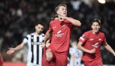 En spiller fra Spartak Moskva feirer etter å ha scoret mål i en fotballkamp. Flere motspillere står i bakgrunnen, og kampen foregår på en stadion med tilskuere. Spillerne er iført røde og svarte drakter. Det er kveldstid, med lys fra stadionbelysningen tydelig synlig.