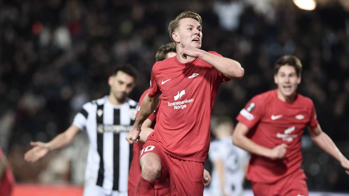 En spiller fra Spartak Moskva feirer etter å ha scoret mål i en fotballkamp. Flere motspillere står i bakgrunnen, og kampen foregår på en stadion med tilskuere. Spillerne er iført røde og svarte drakter. Det er kveldstid, med lys fra stadionbelysningen tydelig synlig.