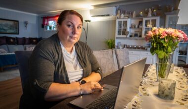 En kvinne sitter ved et bord med en laptop, iført en grå genser. Rundt henne er stoler med mønstrete trekk, og en vase med blomster står på bordet. I bakgrunnen er det en hylle med dekorative gjenstander og planter. Rommet har et varmt og koselig preg med naturlig lys.