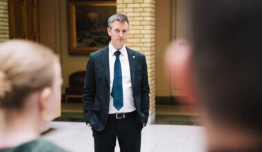 En mann i dress står foran to personer i en innendørs setting. Bakgrunnen viser gule mursteinsvegger og et maleri. Mannen har hendene i lommene og ser mot den som står nærmest. De to personene i forgrunnen er delvis synlige.