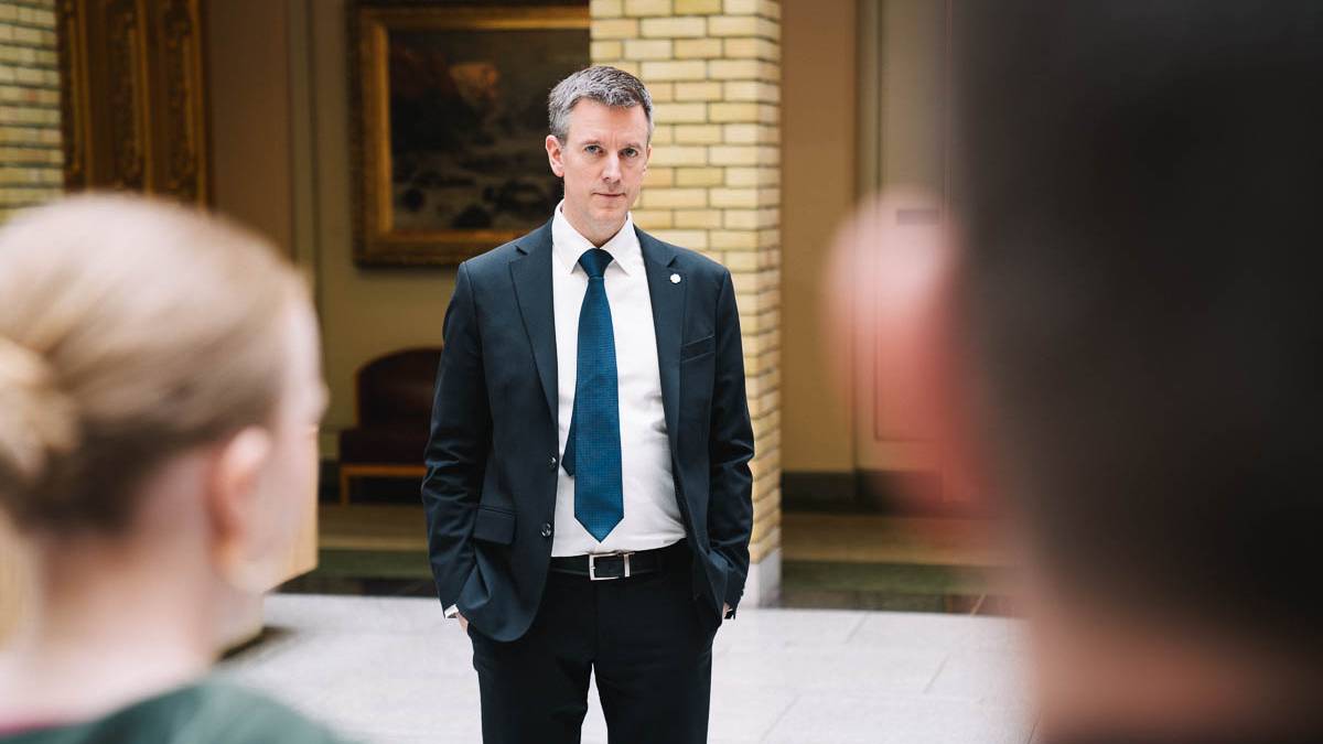 En mann i dress står foran to personer i en innendørs setting. Bakgrunnen viser gule mursteinsvegger og et maleri. Mannen har hendene i lommene og ser mot den som står nærmest. De to personene i forgrunnen er delvis synlige.