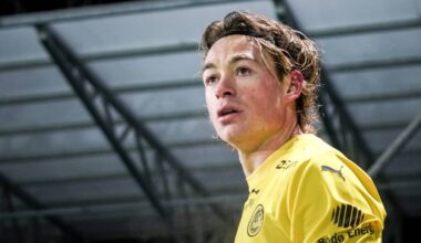 En fotballspiller i gul drakt står på banen. Drakten har merker fra sponsorer. Bakgrunnen er uskarp, men antyder en stadion. Spilleren ser alvorlig ut, muligens etter en kamp.