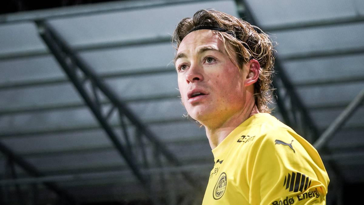 En fotballspiller i gul drakt står på banen. Drakten har merker fra sponsorer. Bakgrunnen er uskarp, men antyder en stadion. Spilleren ser alvorlig ut, muligens etter en kamp.