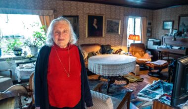 En kvinne med bølgete hvitt hår står i stuen. Hun har på seg en rød genser med lange ermer. Bakgrunnen viser en koselig stue med en stor sofa, et bord med duk og flere planter. Lys fra et vindu og lampe gir en varm atmosfære.