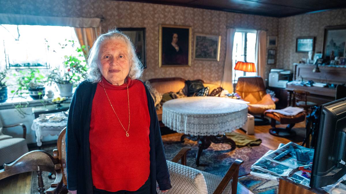 En kvinne med bølgete hvitt hår står i stuen. Hun har på seg en rød genser med lange ermer. Bakgrunnen viser en koselig stue med en stor sofa, et bord med duk og flere planter. Lys fra et vindu og lampe gir en varm atmosfære.