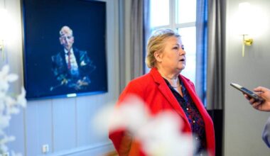 En kvinne i rød jakke står i et rom og prater med en person som holder en mobiltelefon. I bakgrunnen henger et portrett av en mann. Rommet har gardiner og vegger i en lys farge. Det er også blomster i forkant av bildet.