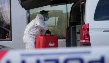 En person i hvit beskyttelsesdrakt bærer en rød kasse ut av en bil. Kassen har teksten "Leica" på seg. Omgivelsene inkluderer en bygning med vinduer og gardiner, samt en politisperring i forgrunnen. Hendelsen skjer i dagslys, og det er klart at personen arbeider med utstyr. (Bildebeskrivelsen er laget av en KI-tjeneste)