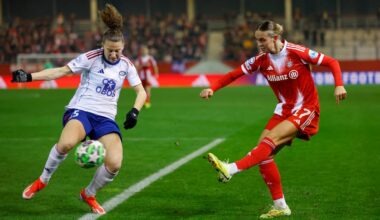 To personer i fotballsituasjon på en bane. Den ene spilleren, iført en hvit drakt med blå detaljer, prøver å blokkere ballen fra en motspiller iført rød drakt. Begge spillere viser tydelig fysisk innsats. Stadionet i bakgrunnen er fylt med tilskuere. (Bildebeskrivelsen er laget av en KI-tjeneste)