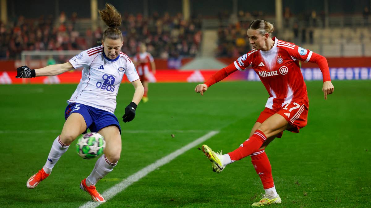 To personer i fotballsituasjon på en bane. Den ene spilleren, iført en hvit drakt med blå detaljer, prøver å blokkere ballen fra en motspiller iført rød drakt. Begge spillere viser tydelig fysisk innsats. Stadionet i bakgrunnen er fylt med tilskuere. (Bildebeskrivelsen er laget av en KI-tjeneste)