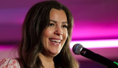 En kvinne holder en mikrofon og synger på en scene med rosa belysning i bakgrunnen. Det er en intim setting som tyder på en live opptreden. Hun har langt, bølgete hår og en bluse med blomsterdesign. Omgivelsene er ukjente, men fargene skaper en livlig atmosfære.