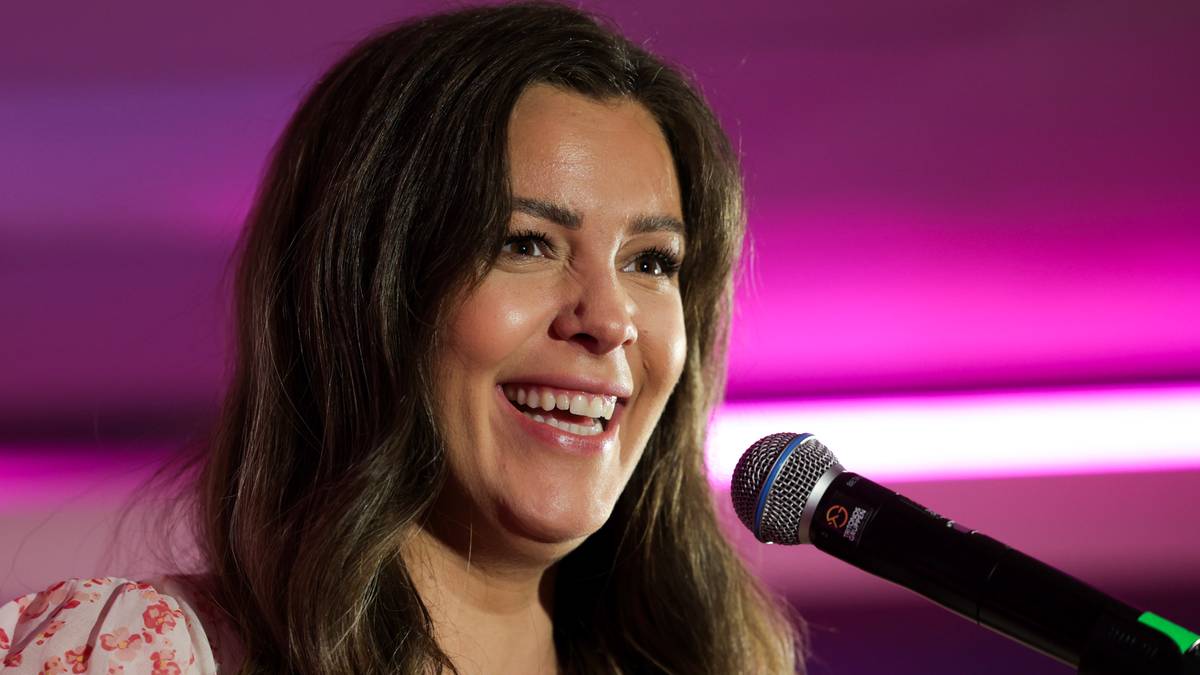 En kvinne holder en mikrofon og synger på en scene med rosa belysning i bakgrunnen. Det er en intim setting som tyder på en live opptreden. Hun har langt, bølgete hår og en bluse med blomsterdesign. Omgivelsene er ukjente, men fargene skaper en livlig atmosfære.