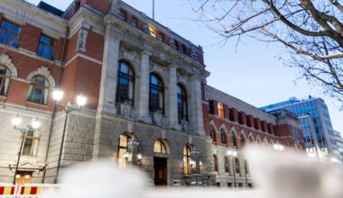 Bygningen av bildet er en historisk struktur med detaljer i nyrenessansestil, beliggende i en urban setting. Det er synlige gatelys og trær i forgrunnen, og himmelen er farget i blå nyanser som signaliserer skumring. Det er også sikkerhetsbarrierer i området, muligens som følge av pågående arbeid eller arrangementer. Bygningens fasade viser detaljer som er typiske for tidsepoken. (Bildebeskrivelsen er laget av en KI-tjeneste)