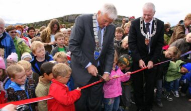 To menn i dress kutter et rødt bånd sammen med en gruppe barn under en åpningsseremoni. Barn i forskjellige klær står rundt dem, noen ser spent på hendelsen. Det er en uklar bakgrunn med grønn natur. Kameraer er synlige, noe som tyder på mediedekning av arrangementet. (Bildebeskrivelsen er laget av en KI-tjeneste)