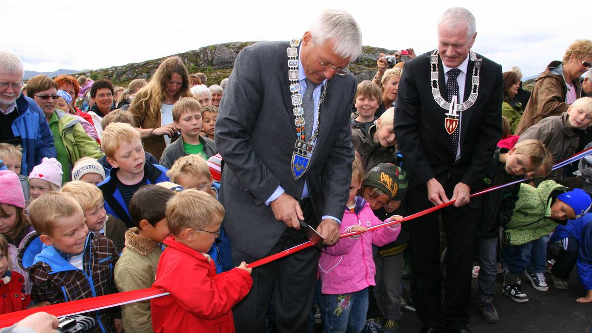 To menn i dress kutter et rødt bånd sammen med en gruppe barn under en åpningsseremoni. Barn i forskjellige klær står rundt dem, noen ser spent på hendelsen. Det er en uklar bakgrunn med grønn natur. Kameraer er synlige, noe som tyder på mediedekning av arrangementet. (Bildebeskrivelsen er laget av en KI-tjeneste)