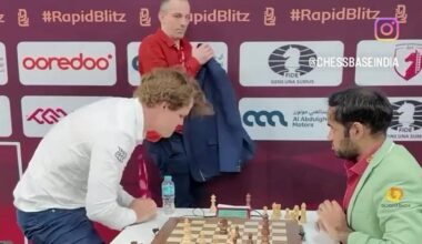 To personer spiller sjakk ved et bord med sjakkbrikker. Den ene spilleren ser ut til å uttrykke frustrasjon ved å slå ned i bordet. Omgivelsene inkluderer et arrangement i Qatar, med elementi av sjakkturneringen synlige i bakgrunnen. Det er et banner med logoer og sponsorer bak dem. (Bildebeskrivelsen er laget av en KI-tjeneste)