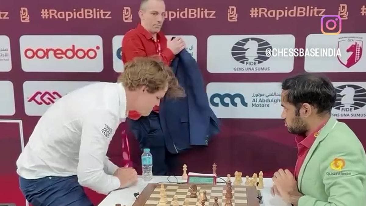 To personer spiller sjakk ved et bord med sjakkbrikker. Den ene spilleren ser ut til å uttrykke frustrasjon ved å slå ned i bordet. Omgivelsene inkluderer et arrangement i Qatar, med elementi av sjakkturneringen synlige i bakgrunnen. Det er et banner med logoer og sponsorer bak dem. (Bildebeskrivelsen er laget av en KI-tjeneste)