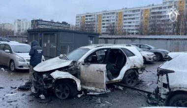 Et ødelagt bilvrak står i en parkeringsplass, med det meste av frontpartiet bortknekt. En person i uniform undersøker skadeomfanget. Bakgrunnen viser flere parkerte biler og bolighus. Det er overskyet vær, og området ser fuktig ut. (Bildebeskrivelsen er laget av en KI-tjeneste)