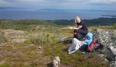 To personer og en hund sitter på en stein i en fjellandskap. Den ene personen holder en matbit, mens den andre er iført en hettegenser. Bakgrunnen viser et vidstrakt, naturlig landskap med skog og fjord. Skyene henger lavt på himmelen, og det ser ut til å være overskyet vær.