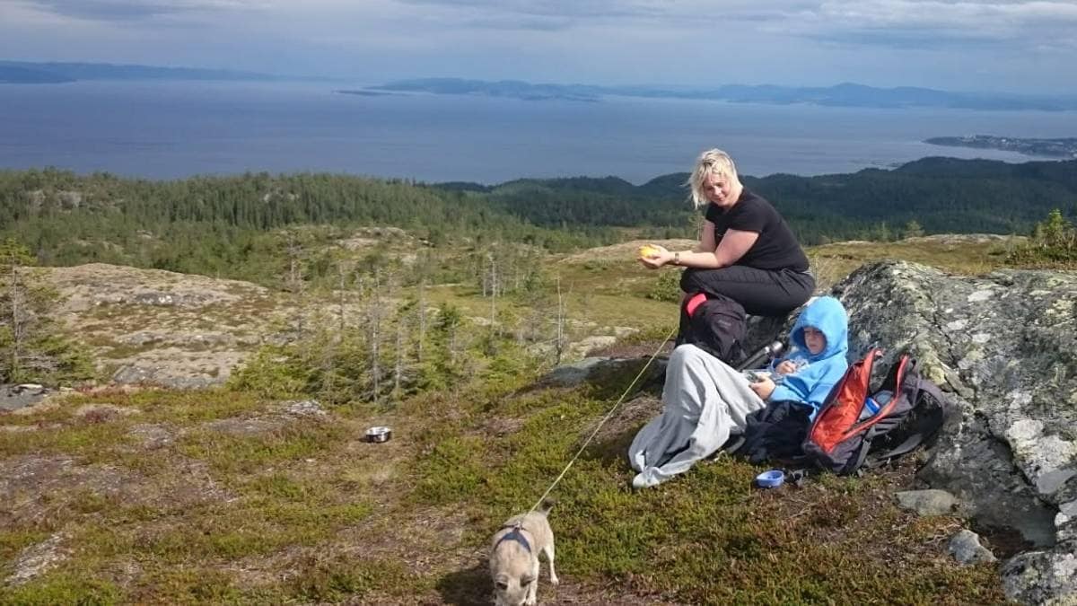 To personer og en hund sitter på en stein i en fjellandskap. Den ene personen holder en matbit, mens den andre er iført en hettegenser. Bakgrunnen viser et vidstrakt, naturlig landskap med skog og fjord. Skyene henger lavt på himmelen, og det ser ut til å være overskyet vær.