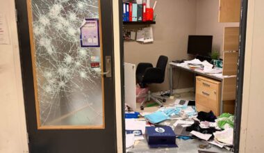 Bilde av en ødelagt dør med sprekker, som leder inn til et rotete kontor. Inne i kontoret er det papirer og gjenstander spredt utover gulvet. En svart kontorstol og et skrivebord med en datamaskin er synlig blant rotet. Omgivelsene er nakne, med grå vegger og synlige rør i taket.