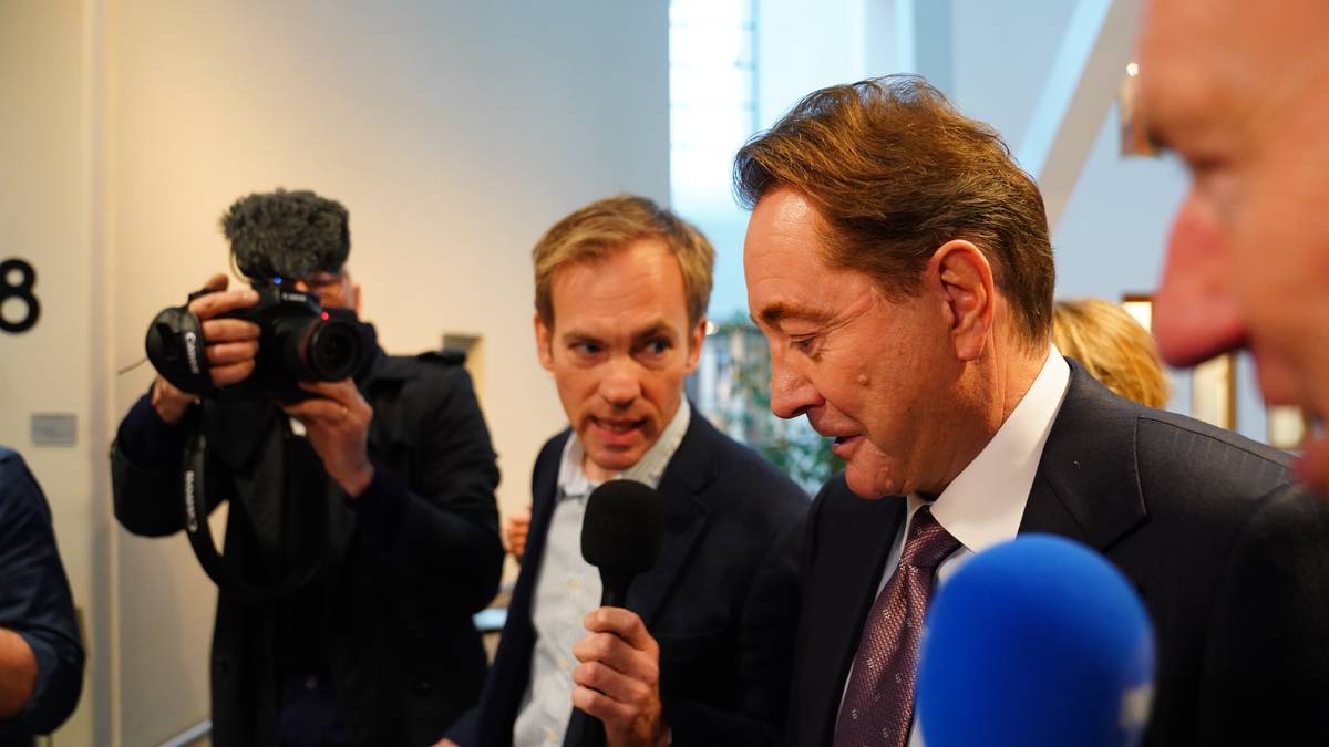 En gruppe mennesker står samlet i et rom. En person holder en mikrofon, mens en annen er iført en kamerabeskyttelse og tar bilder. Det er et moderne interiør med naturlig lys som strømmer inn fra vinduene. Flere personer i bakgrunnen kan sees, og stemningen virker hektisk.