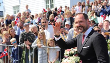 En mann i mørk dress vinker til publikum med buketter i hendene. Bak ham står en stor folkemengde som holder flagg og mobiltelefoner. Omgivelsene inkluderer en hvit bygning og en klar blå himmel. Arrangementet ser ut til å være en feiring eller markering. (Bildebeskrivelsen er laget av en KI-tjeneste)