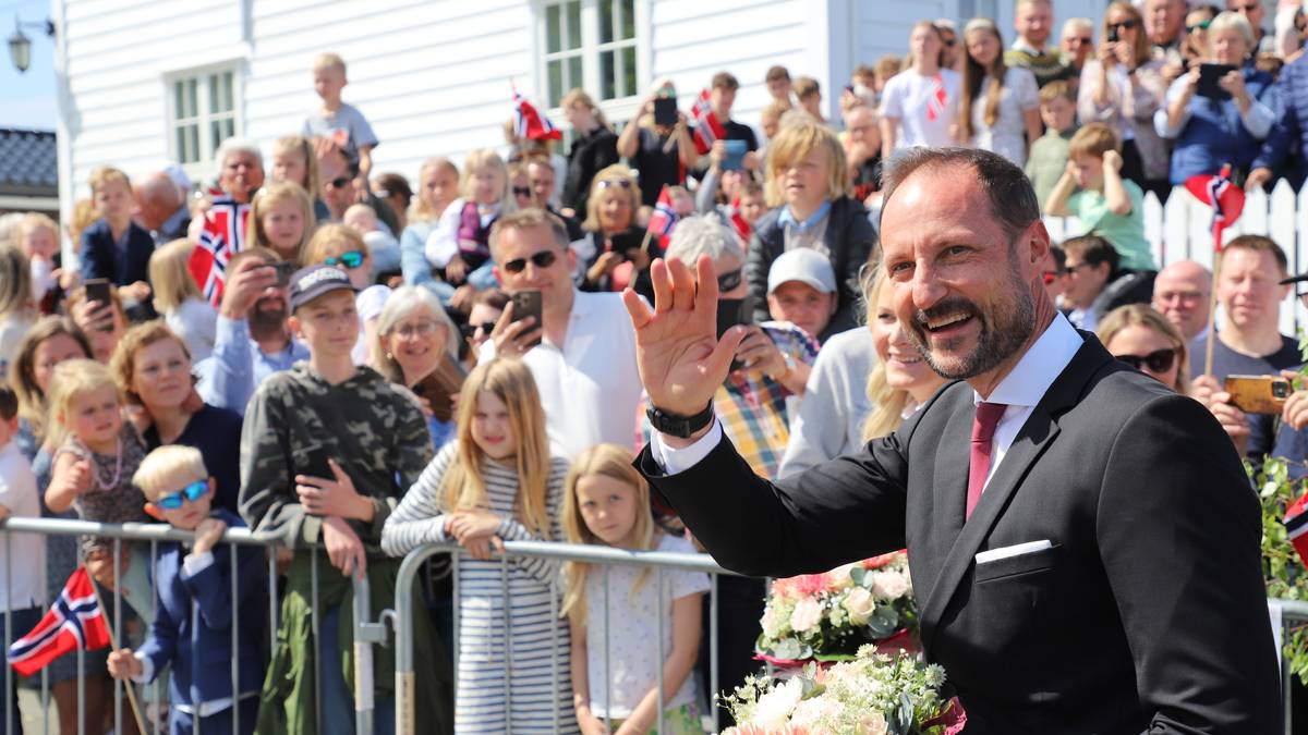 En mann i mørk dress vinker til publikum med buketter i hendene. Bak ham står en stor folkemengde som holder flagg og mobiltelefoner. Omgivelsene inkluderer en hvit bygning og en klar blå himmel. Arrangementet ser ut til å være en feiring eller markering. (Bildebeskrivelsen er laget av en KI-tjeneste)