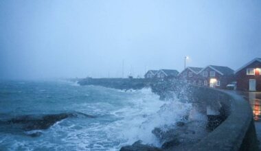 Uvær i Bodø i Nordland. Bølgene slår inn mot land over moloen i Bodø og skyller over røde sjøhus. I bakgrunn synes byen i snøvær.