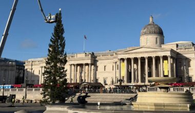 En arbeider i en heis monterer en stjerne på toppen av et juletre i Trafalgar Square. Bakgrunnen viser National Gallery, med en stor kuppel og søyler. Flere personer i hi-viz klær kan sees arbeide på plassen. Klar blå himmel og julemarked er synlig til venstre.
