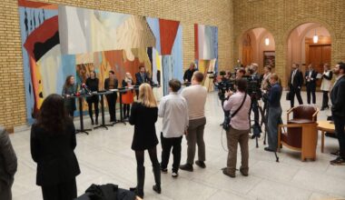 En pressekonferanse finner sted i en romslig sal med kunstneriske vegger. Flere personer står bak et bord, hvor en kvinne taler. Journalister og kamerafolk er samlet foran, og noen sitter i stoler på siden. Omgivelsene består av murvegger og storartede detaljer.