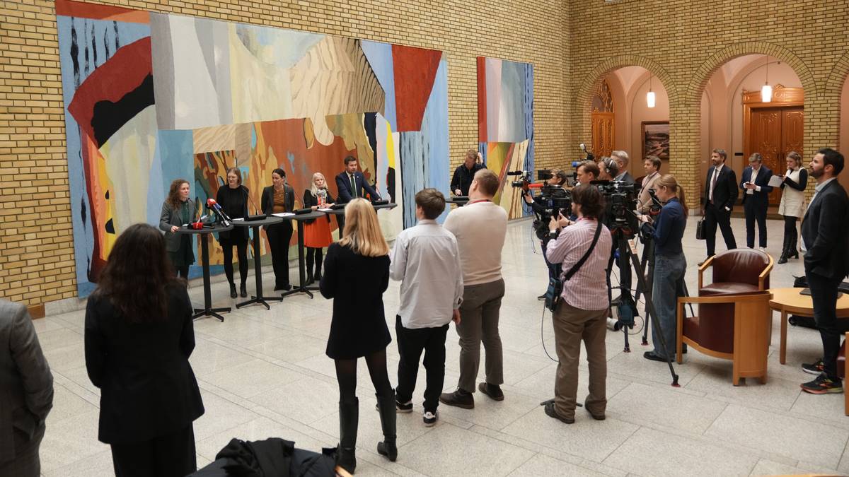 En pressekonferanse finner sted i en romslig sal med kunstneriske vegger. Flere personer står bak et bord, hvor en kvinne taler. Journalister og kamerafolk er samlet foran, og noen sitter i stoler på siden. Omgivelsene består av murvegger og storartede detaljer.