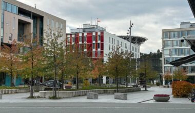 Bilde viser et bylandskap med moderne bygninger i forskjellige farger og former. I sentrum er det en åpen plass med trær i høstfarger. Det er biler parkert i nærheten, og en del mennesker synlig i bakgrunnen. En hellikopterplattform er synlig i det fjerne, som gir kontekst til området.