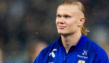 En fotballspiller står i fokus iført en blå jakke med norsk fotballlogo. Det er regnvær, og dråper kan sees på kameravinkelen. Bakgrunnen har uskarpe figurer som indikerer en sportsarena. Spilleren ser mot siden, tydelig konsentrert før en kamp.