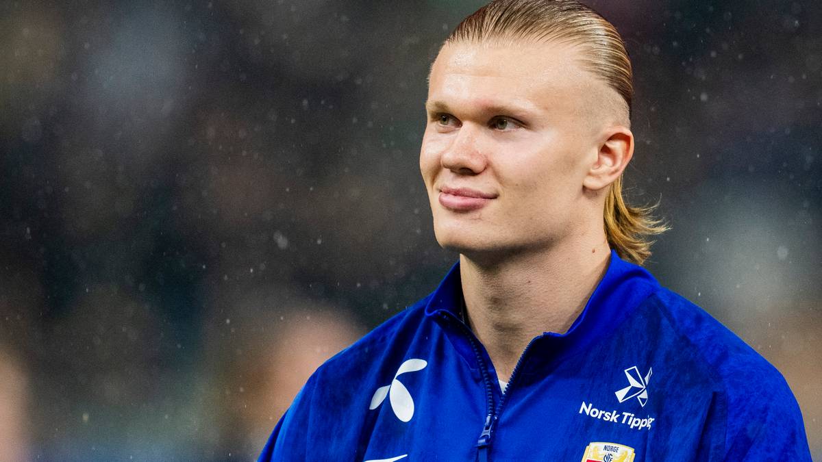 En fotballspiller står i fokus iført en blå jakke med norsk fotballlogo. Det er regnvær, og dråper kan sees på kameravinkelen. Bakgrunnen har uskarpe figurer som indikerer en sportsarena. Spilleren ser mot siden, tydelig konsentrert før en kamp.