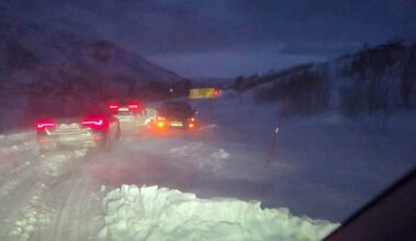 Bildet viser en snødekt vei i mørket, med flere biler som står i kø. I bakgrunnen er det en lastebil og en snøskuter synlig. Veien er omgitt av snøfylte områder og fjell, noe som indikerer vinterforhold. Bildeutsnittet er tatt fra innsiden av en bil, med lysning fra biler som reflekteres i snøen. (Bildebeskrivelsen er laget av en KI-tjeneste)