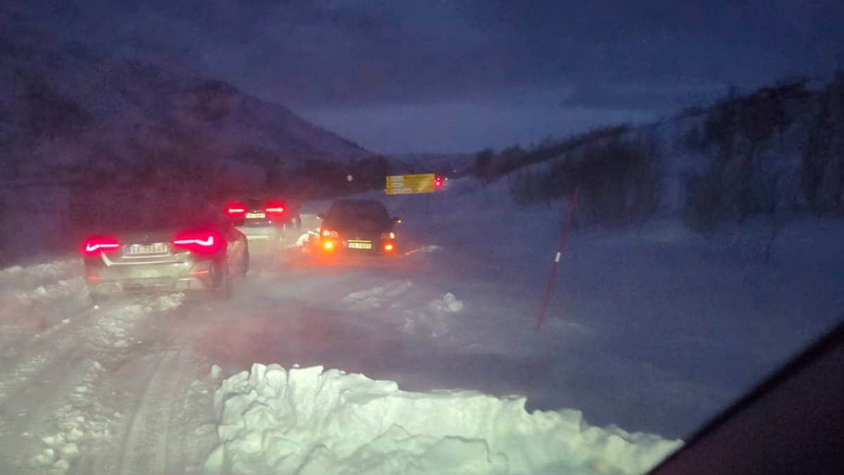 Bildet viser en snødekt vei i mørket, med flere biler som står i kø. I bakgrunnen er det en lastebil og en snøskuter synlig. Veien er omgitt av snøfylte områder og fjell, noe som indikerer vinterforhold. Bildeutsnittet er tatt fra innsiden av en bil, med lysning fra biler som reflekteres i snøen. (Bildebeskrivelsen er laget av en KI-tjeneste)