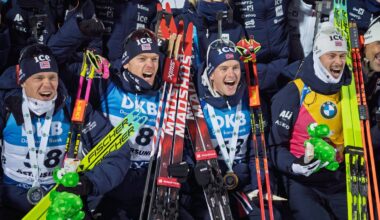 Bildet viser en gruppe langrennsløpere som poserer sammen iført matchende novemberklær. De holder ski og medaljer, noe som indikerer en prestasjon i løpet av en konkurranse. En utøver holder også en grønn leke i form av en dinosaur. Omgivelsene bærer preg av en sportslig setting, muligens en premieutdeling. (Bildebeskrivelsen er laget av en KI-tjeneste)