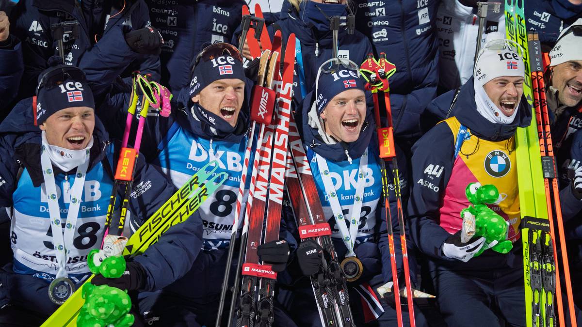 Bildet viser en gruppe langrennsløpere som poserer sammen iført matchende novemberklær. De holder ski og medaljer, noe som indikerer en prestasjon i løpet av en konkurranse. En utøver holder også en grønn leke i form av en dinosaur. Omgivelsene bærer preg av en sportslig setting, muligens en premieutdeling. (Bildebeskrivelsen er laget av en KI-tjeneste)