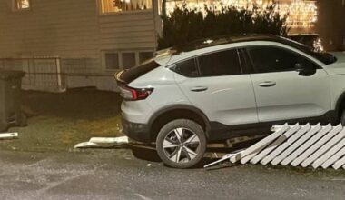 Et grått SUV kjøretøy står på en vei foran en bolig med vinduer lysende i natten. En del av et gjerde ligger på bakken ved siden av bilen, som antyder at det har vært en kollisjon. Boligen har julelys hengende, og det er mørkt i omgivelsene. Ingen personer er synlige i bildet. (Bildebeskrivelsen er laget av en KI-tjeneste)