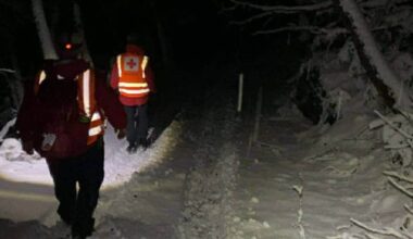 To redningspersonell i refleksvester går langs en snødekt sti om natten. Omgivelsene er preget av mørke, trær med snø og en smal vei. De bærer lykter for å lyse opp stien. Bildet uttrykker en akutt situasjon med fokus på redningsarbeid.