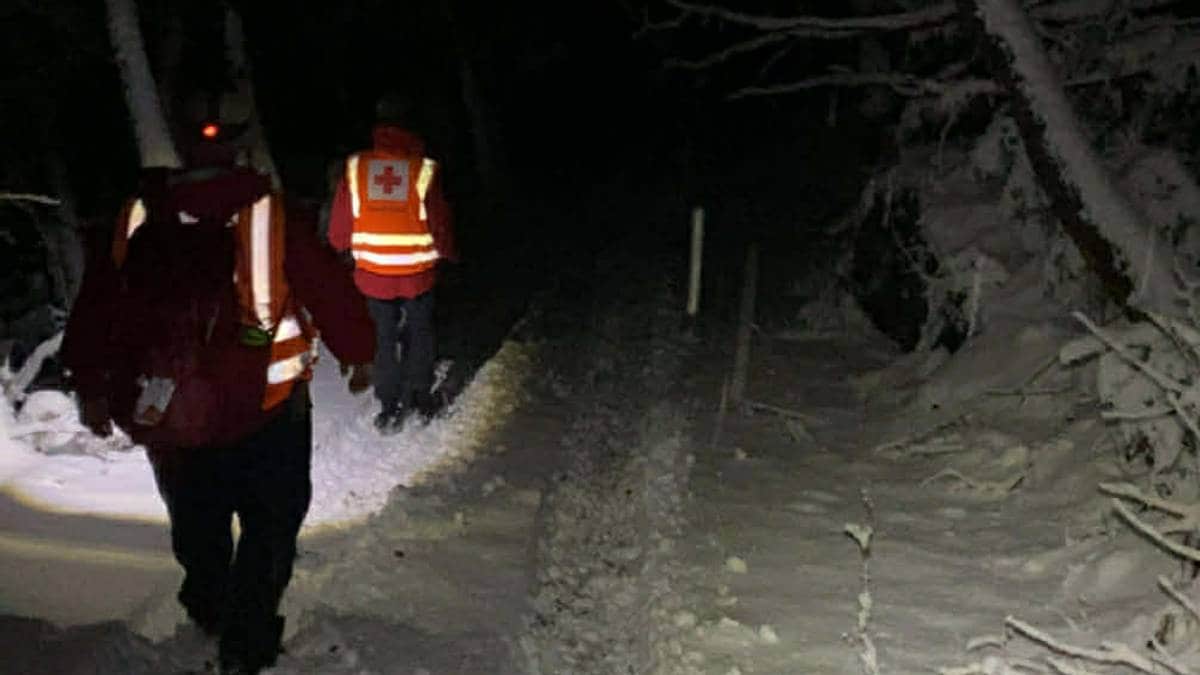 To redningspersonell i refleksvester går langs en snødekt sti om natten. Omgivelsene er preget av mørke, trær med snø og en smal vei. De bærer lykter for å lyse opp stien. Bildet uttrykker en akutt situasjon med fokus på redningsarbeid.