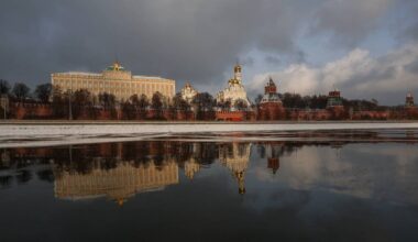 Bildet viser Kreml i Moskva, med den røde muren og tårnene tydelig i front. Bakgrunnen inkluderer en stor bygning med gullkupler og moderne detaljer. Refleksjonen av bygningene i elven gir et visuelt inntrykk av både struktur og natur. Himlen er delvis skyet, noe som gir en dramatisk atmosfære over scenen. (Bildebeskrivelsen er laget av en KI-tjeneste)