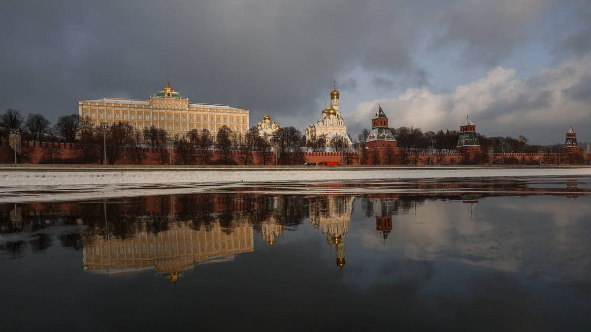 Bildet viser Kreml i Moskva, med den røde muren og tårnene tydelig i front. Bakgrunnen inkluderer en stor bygning med gullkupler og moderne detaljer. Refleksjonen av bygningene i elven gir et visuelt inntrykk av både struktur og natur. Himlen er delvis skyet, noe som gir en dramatisk atmosfære over scenen. (Bildebeskrivelsen er laget av en KI-tjeneste)