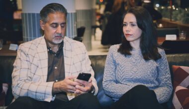 To personer sitter på en sofa i en kafé. Den ene personen holder en smarttelefon, mens den andre sitter stille. Omgivelsen er moderne, med mørke møbler og dempet belysning. Det er flere mennesker i bakgrunnen som samtaler.