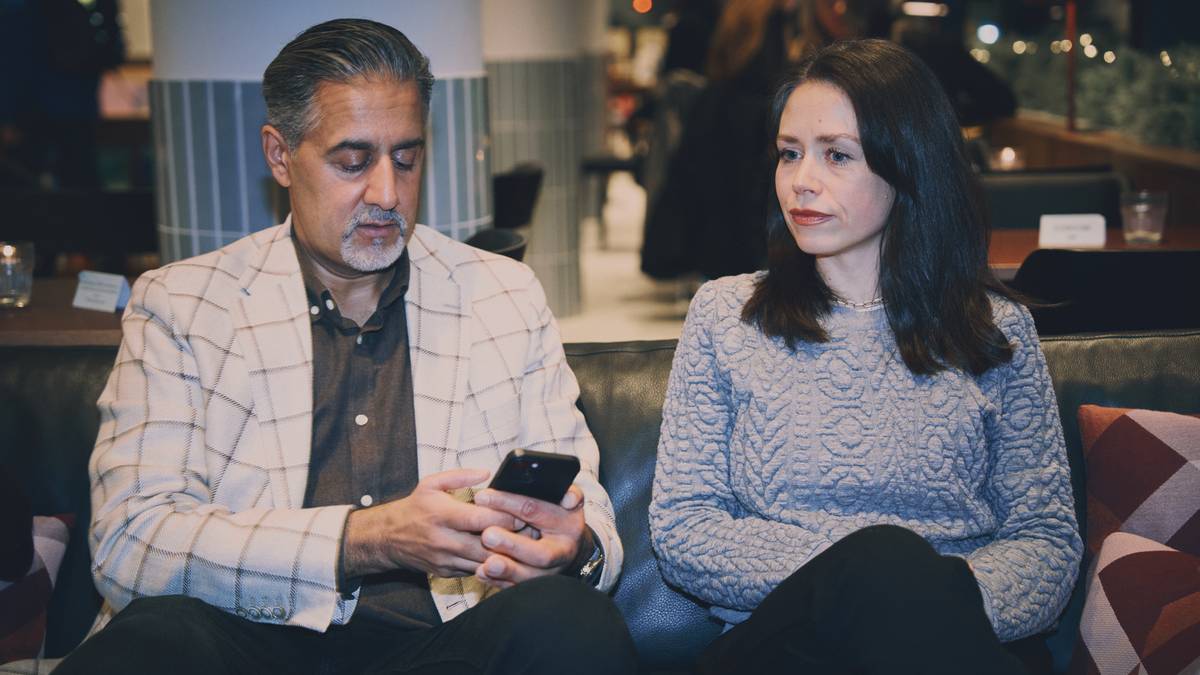 To personer sitter på en sofa i en kafé. Den ene personen holder en smarttelefon, mens den andre sitter stille. Omgivelsen er moderne, med mørke møbler og dempet belysning. Det er flere mennesker i bakgrunnen som samtaler.